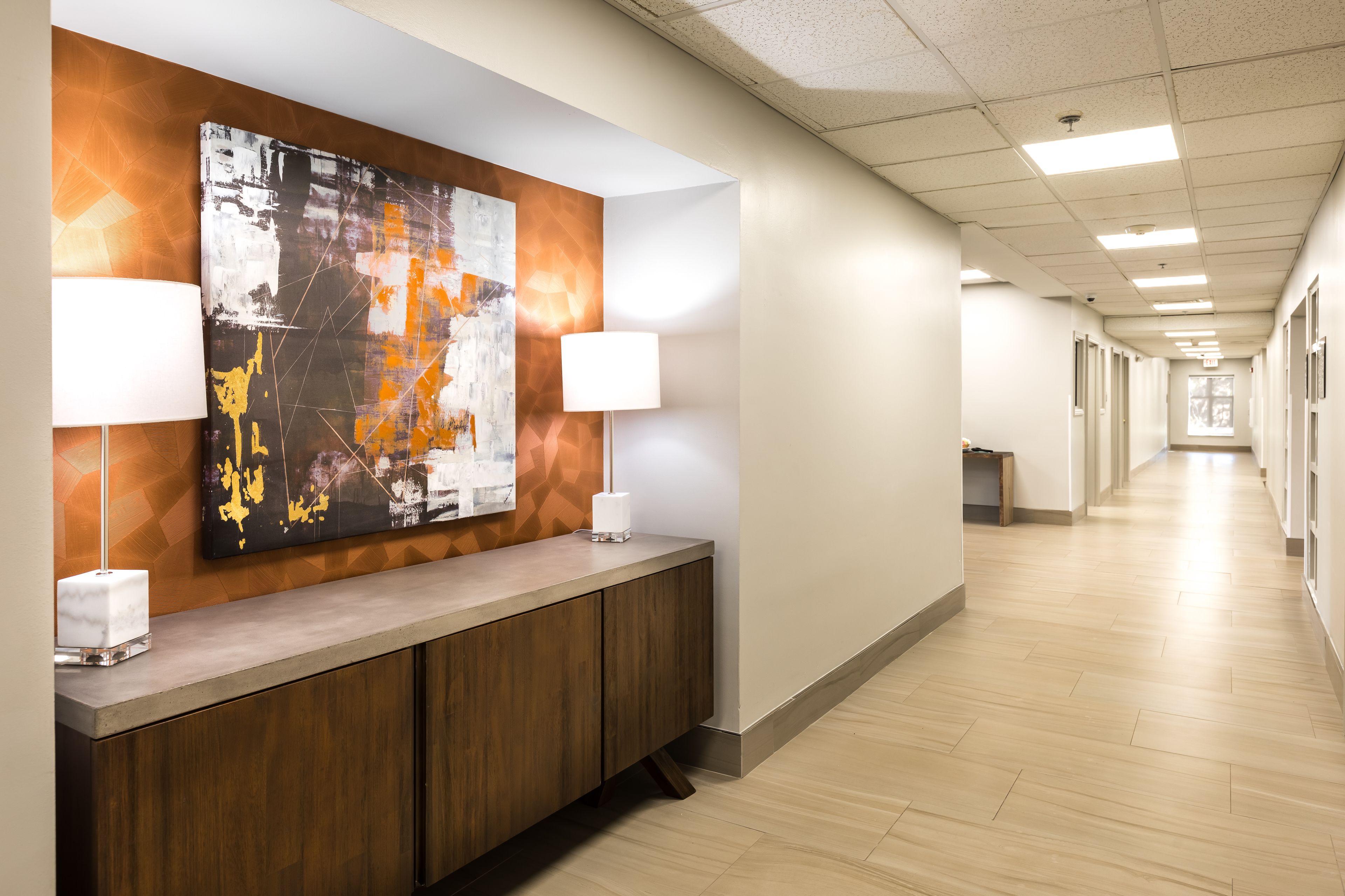 A hotel reception area with a modern design, featuring a long corridor with a reception desk, two lamps, and a large abstract painting on the wall.