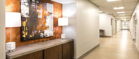 A hotel reception area with a modern design, featuring a long corridor with a reception desk, two lamps, and a large abstract painting on the wall.