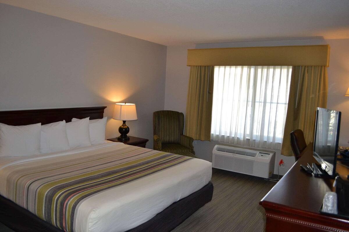 A hotel room with a large bed, a desk with a computer, a chair, a lamp, and an air conditioning unit.