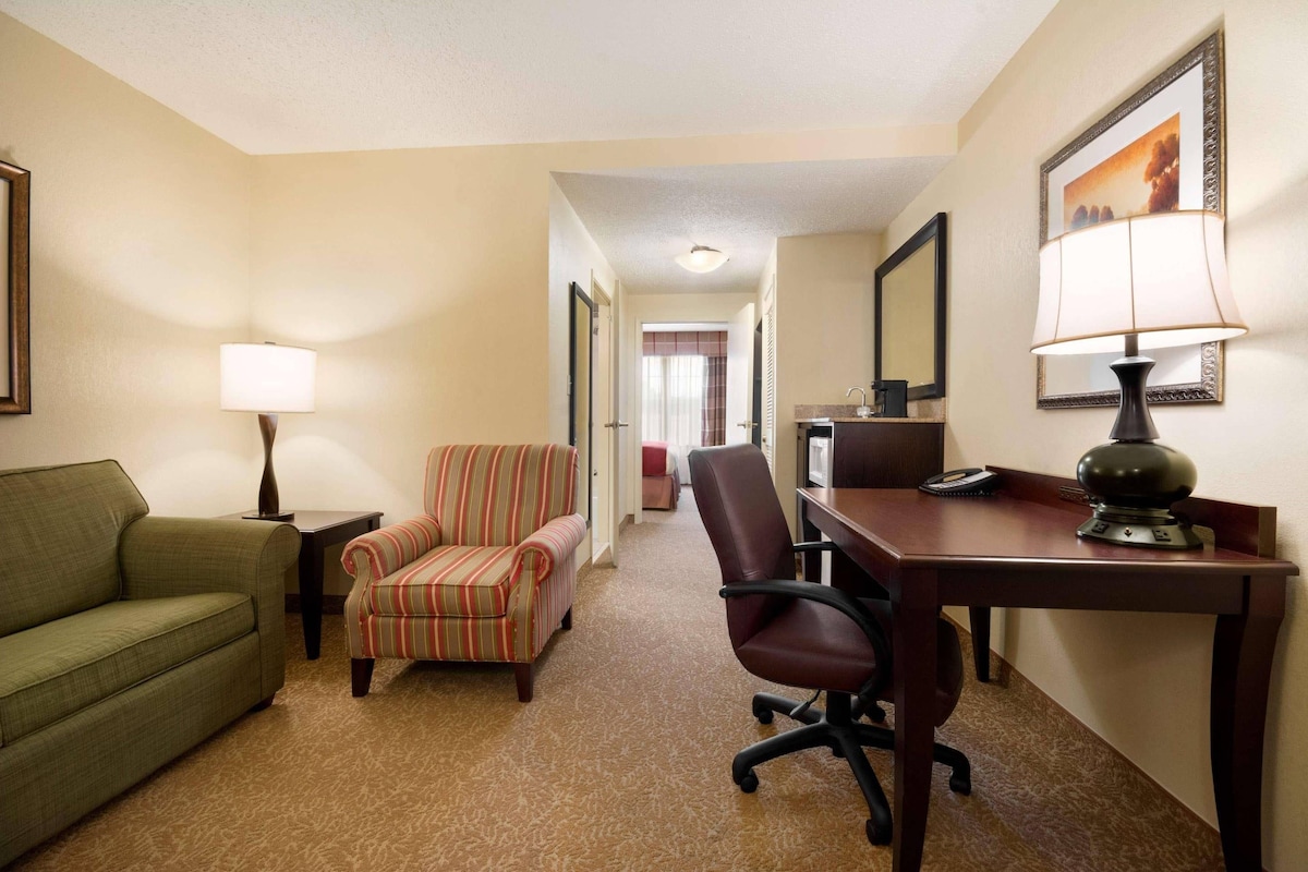 A hotel room with a green sofa, striped armchair, and a desk with a lamp.