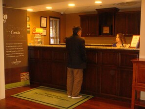 Un hombre está de pie en la recepción de un hotel, con armarios de madera y un certificado enmarcado en la pared.