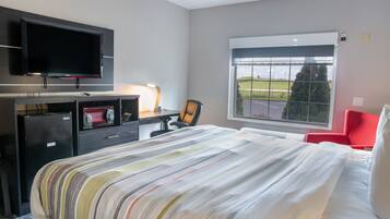 Quarto de hotel com cama, escrivaninha com televisão, cadeira e janela com vista para o exterior.