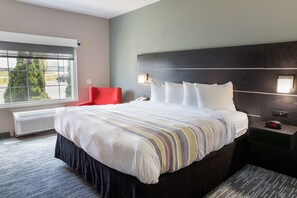 A hotel room with a large bed, a red chair, a nightstand with a clock, and a window with a view of the outside.