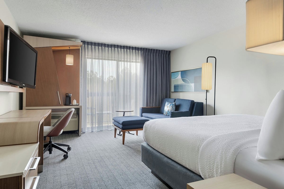 A hotel room with a bed, desk, chair, TV, and a view of the outdoors.