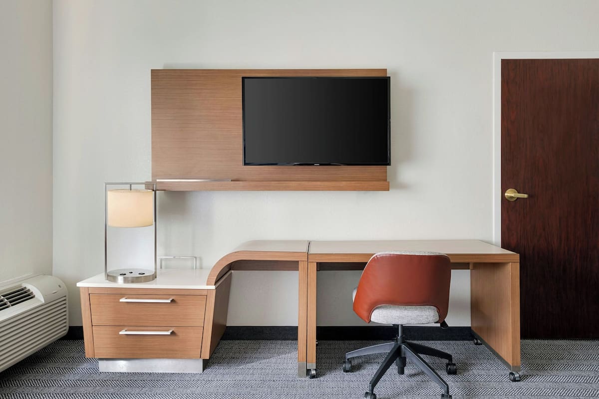 A hotel room with a desk, chair, TV, lamp, and a door.