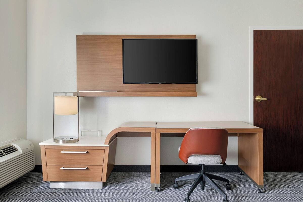 Habitación de hotel con escritorio, silla, televisor, lámpara y puerta.