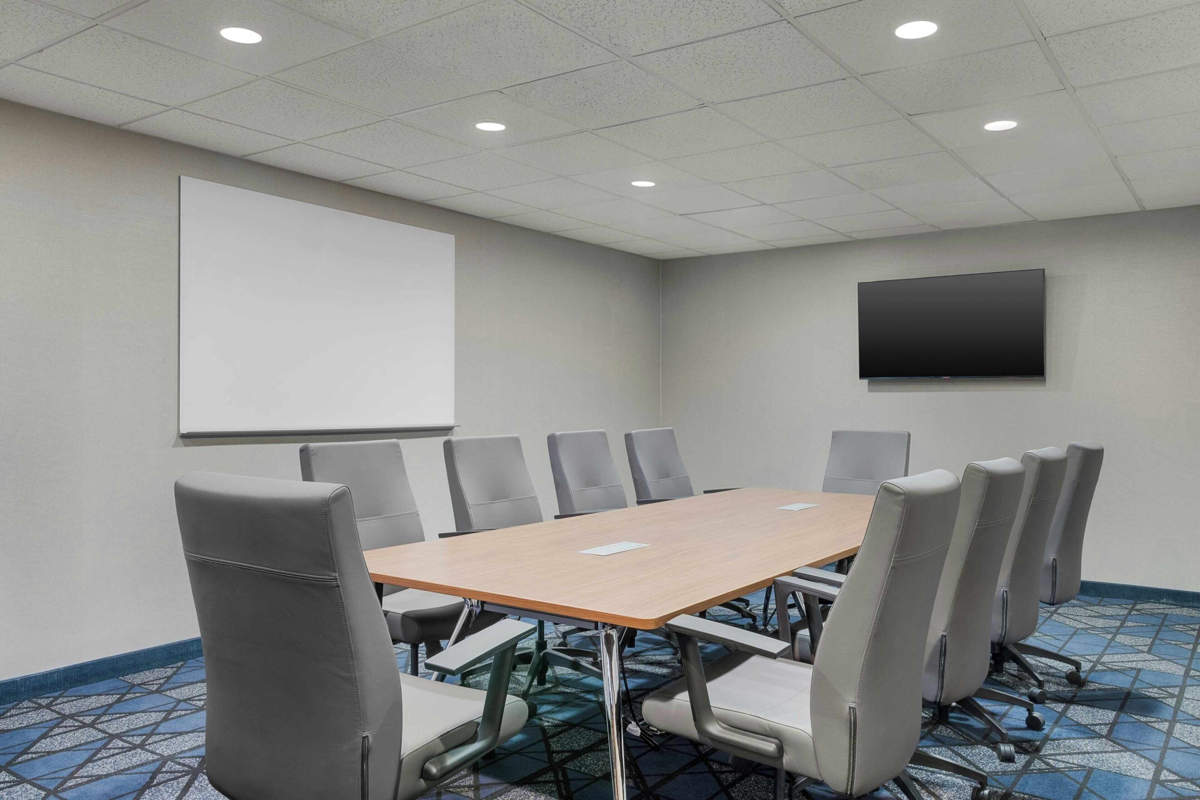 Una sala de conferencias con una mesa grande, sillas grises, una pantalla de proyector, un televisor de pantalla plana y alfombra estampada.