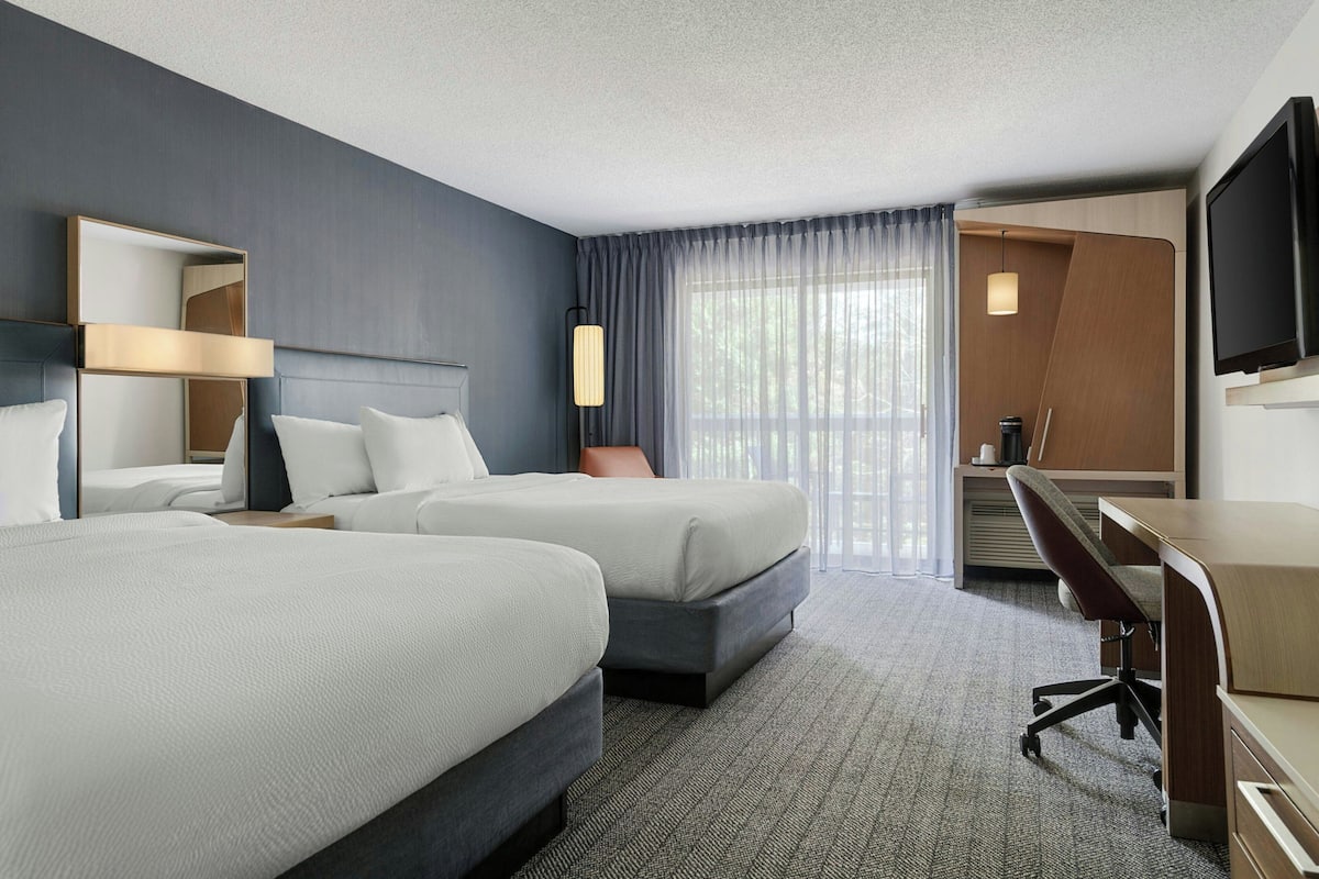 A hotel room with two beds, a desk, a chair, a TV, and a large window with curtains.