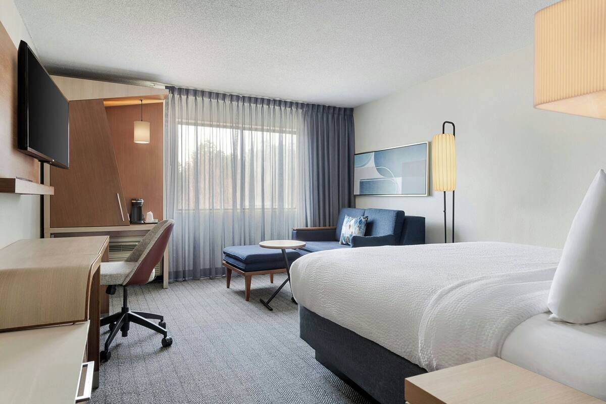 A hotel room with a bed, a desk, a chair, a TV, a lamp, and a window with curtains.