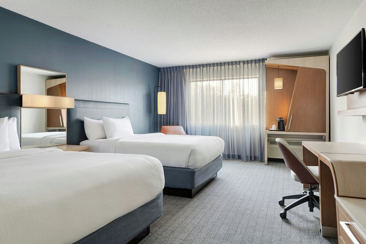 A hotel room with two beds, a desk, a chair, a TV, and a large window with curtains.