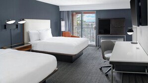 Een hotelkamer met twee bedden, een bureau, een stoel, een televisie en een balkon met uitzicht op de stad.