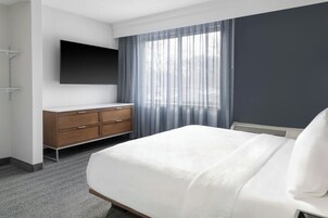 Una habitación de hotel con una cama, un aparador de madera, un televisor de pantalla plana en la pared y un gran ventanal con cortinas translúcidas.