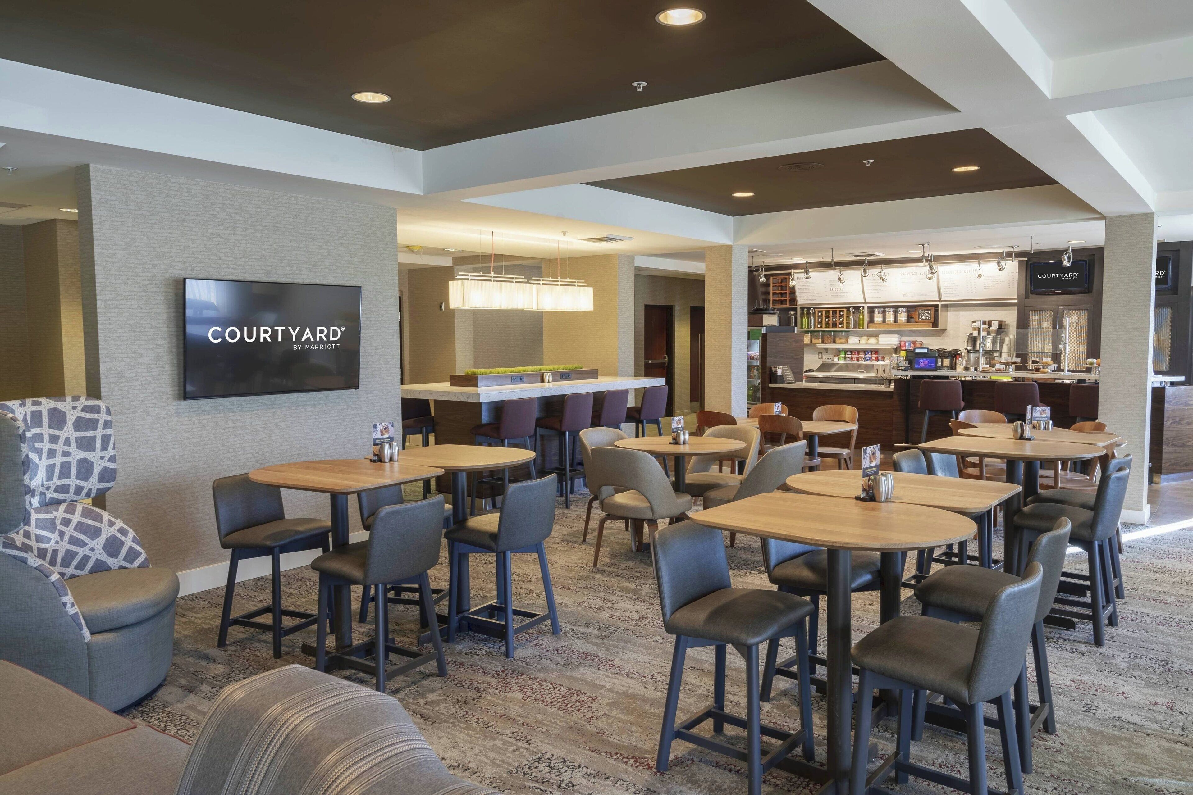 El lobby de un hotel Courtyard by Marriott con varias zonas de estar, un bar y una pantalla de televisión que muestra el nombre del hotel.