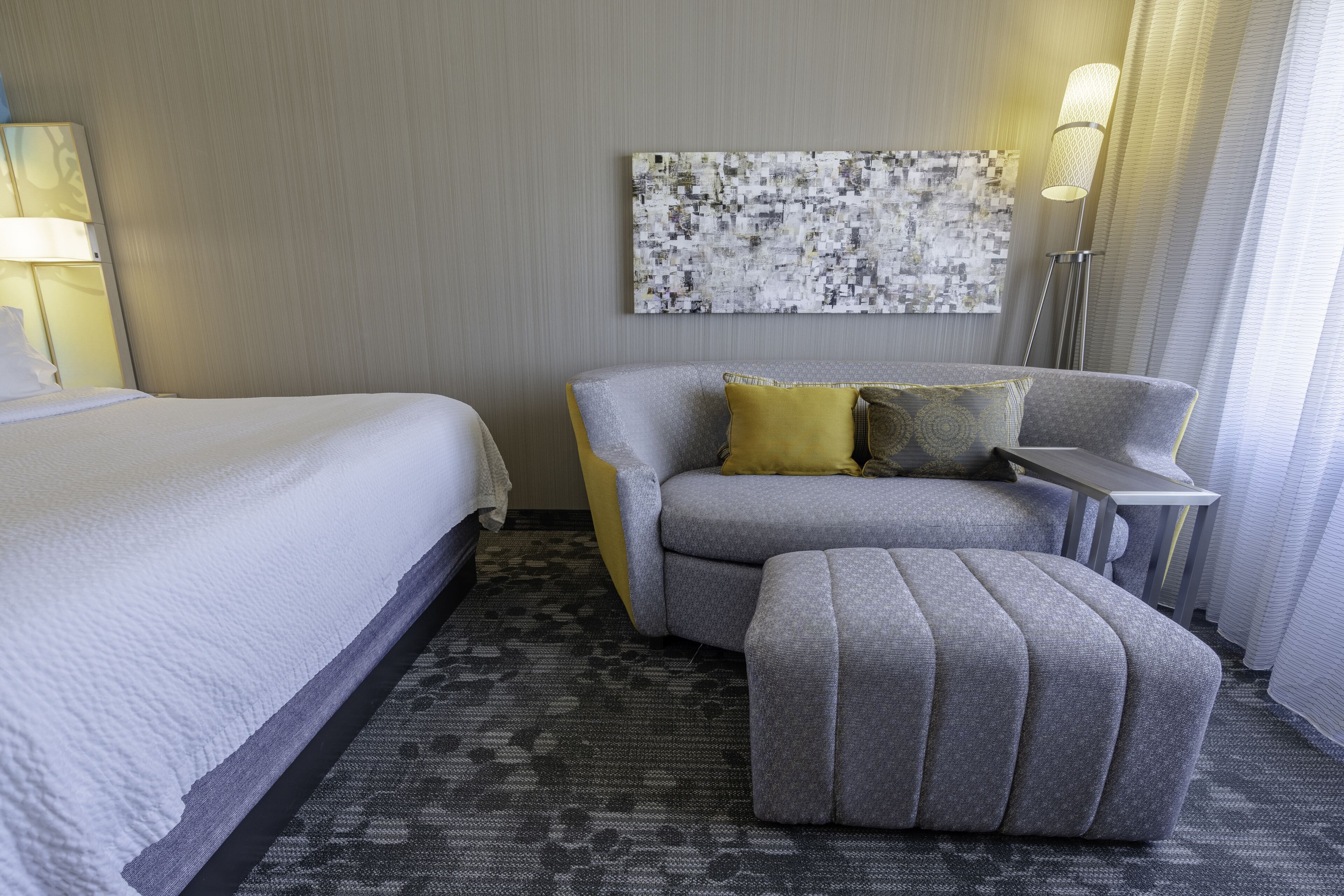 A hotel room with a bed, a sofa, an ottoman, a desk, and a lamp.