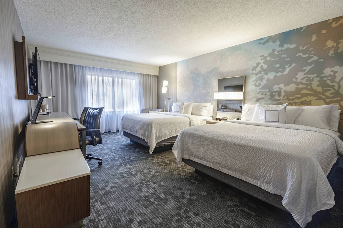 A hotel room with two beds, a desk, a chair, a TV, and a large window with curtains.