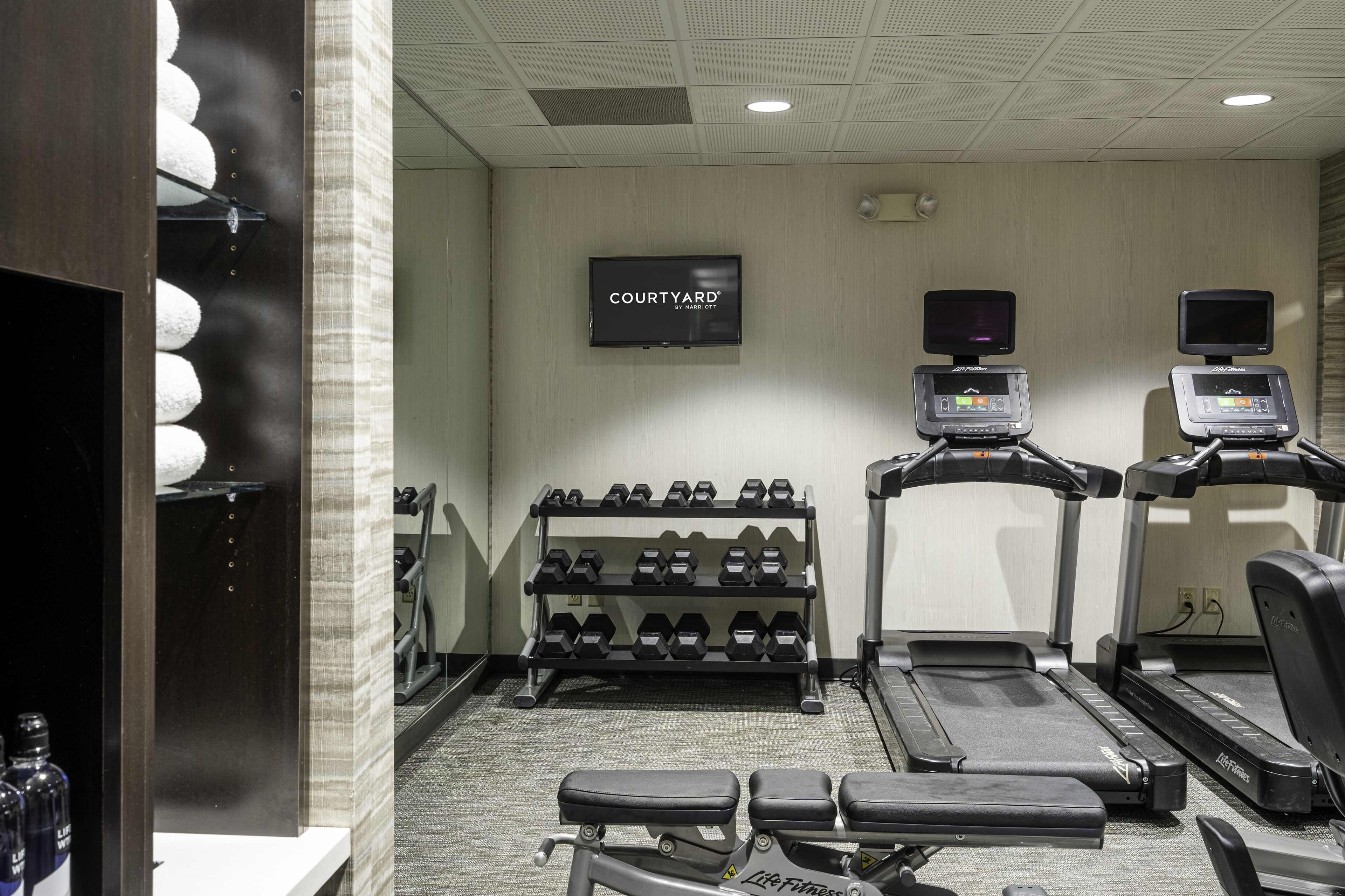 Un gimnasio con cintas de correr, mancuernas y una pantalla de televisión que muestra 'COURTYARD'.
