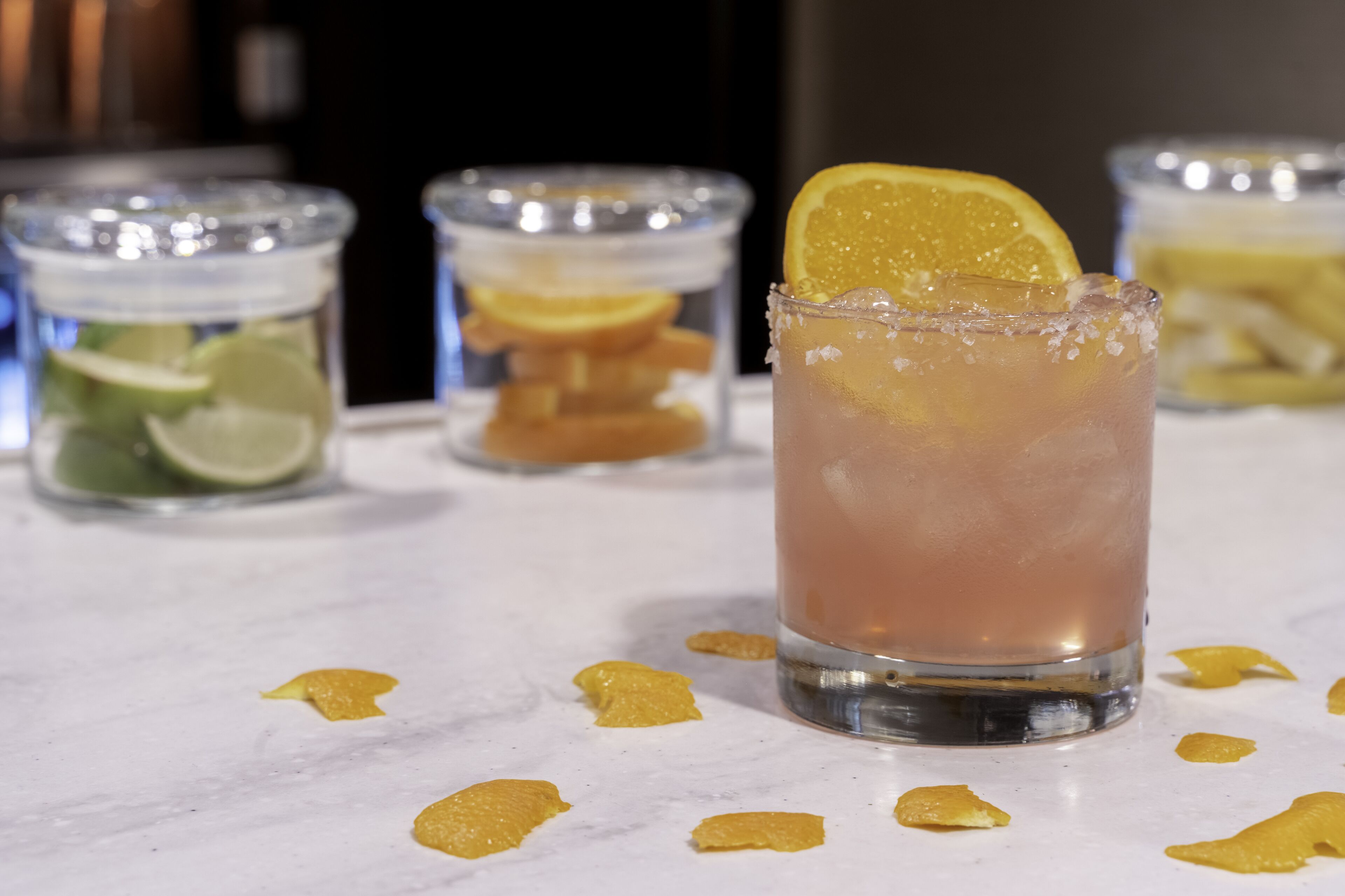 A glass of iced drink with a lemon slice garnish, surrounded by scattered lemon peels on a marble surface.