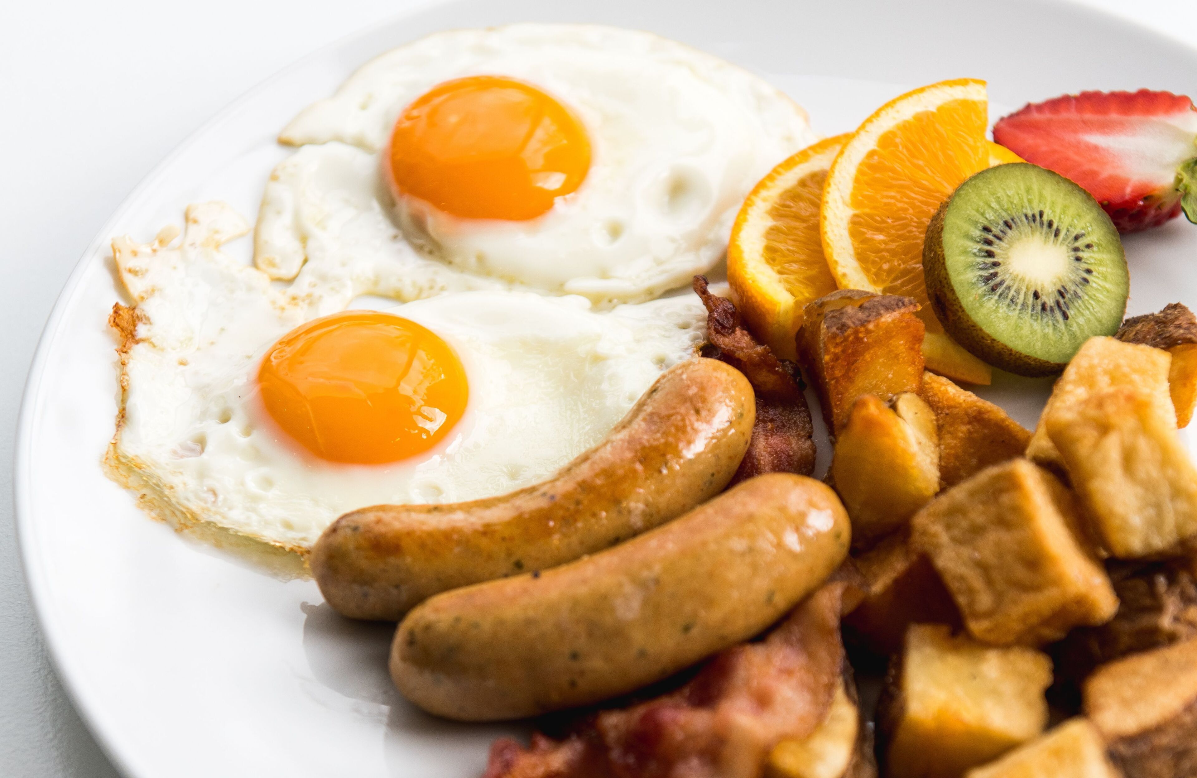 Un piatto con due uova al tegamino, salsiccia, pancetta e frutta fresca, tra cui kiwi e fettine d'arancia.