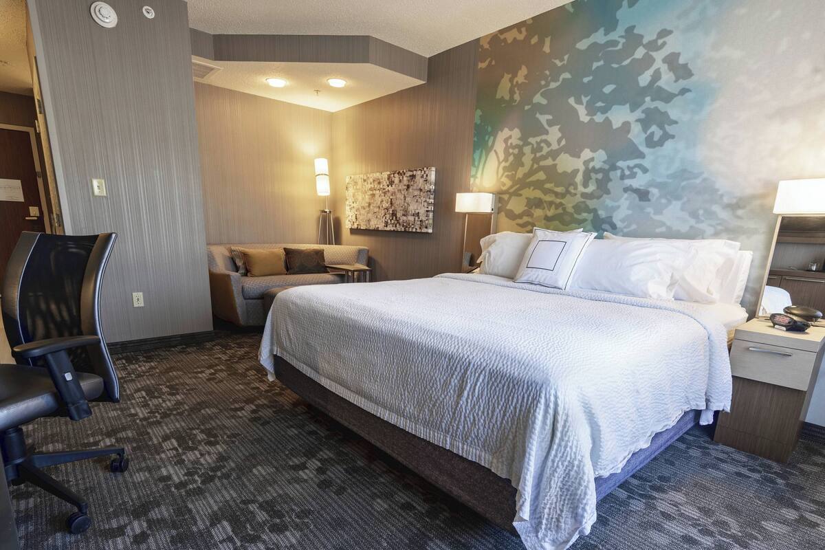 A hotel room with a large bed, a desk, a chair, and a couch. There are two wall-mounted lamps and a ceiling light.