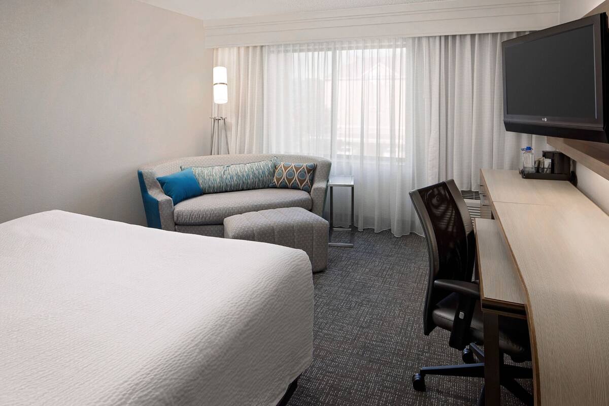 A hotel room with a bed, a sofa, a desk, a chair, a television, and a window with curtains.