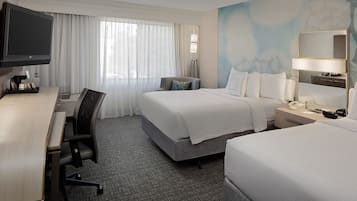 A hotel room with a desk, two beds, a TV, and a large window with curtains.