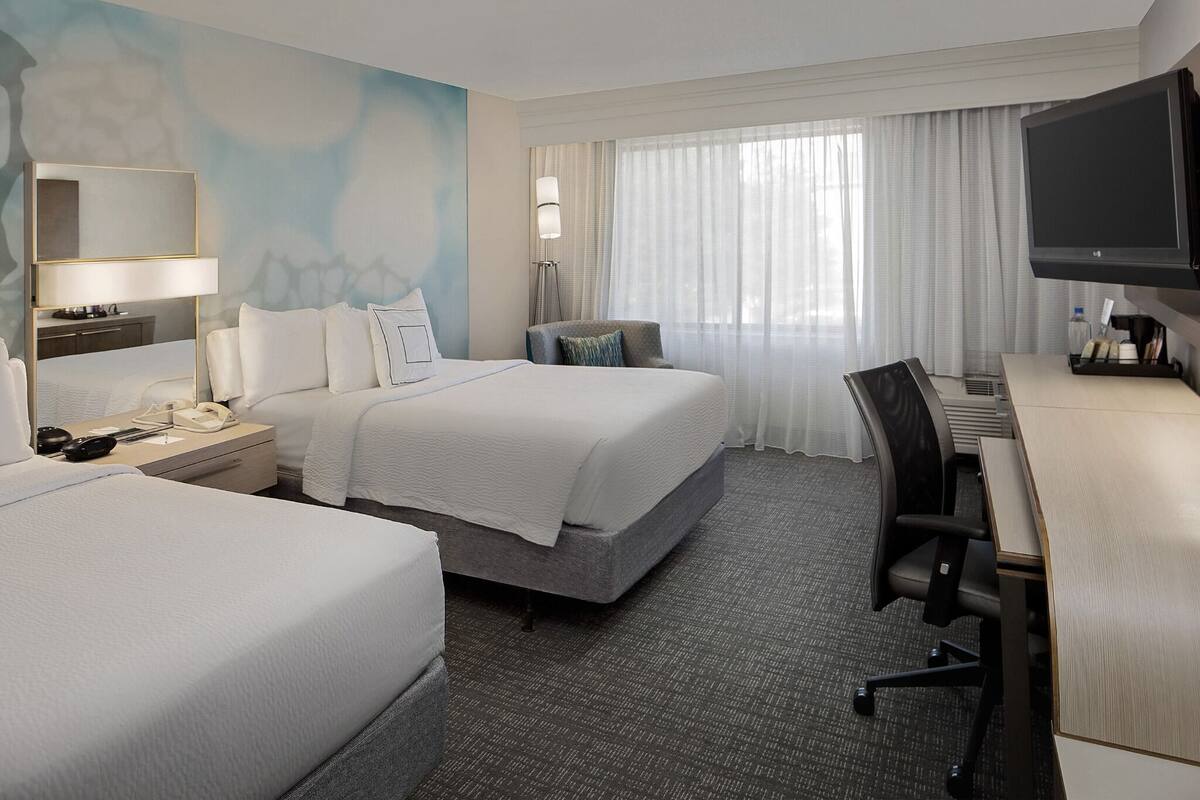 A hotel room with two beds, a desk, a chair, a TV, a lamp, and a window with curtains.