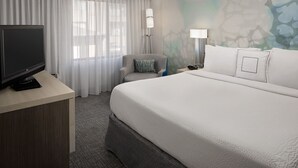 Una habitación de hotel con cama, escritorio con televisor, silla y mesita de noche con lámpara.