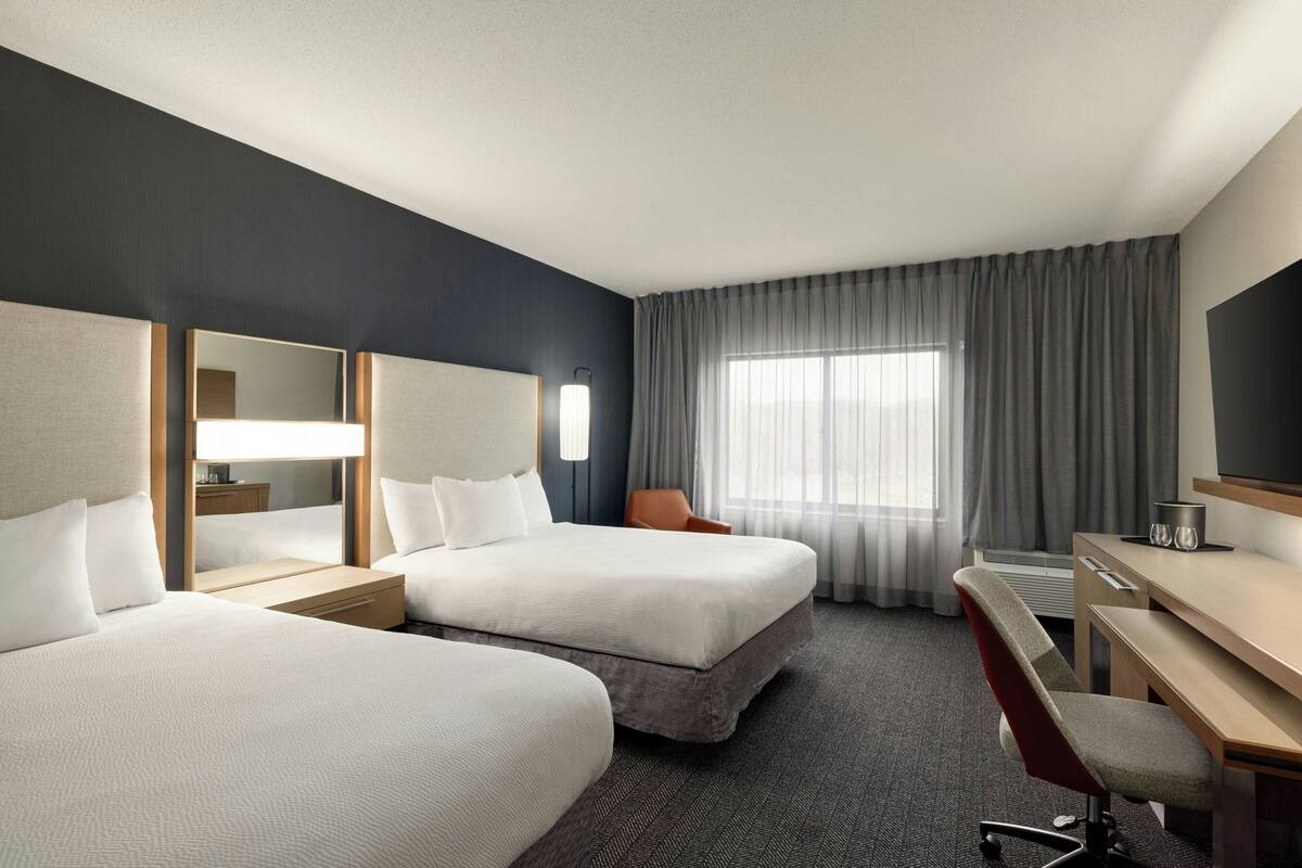 Ein Hotelzimmer mit zwei Betten, einem Schreibtisch, einem Stuhl, einem Spiegel und einem Fenster mit Vorhängen.