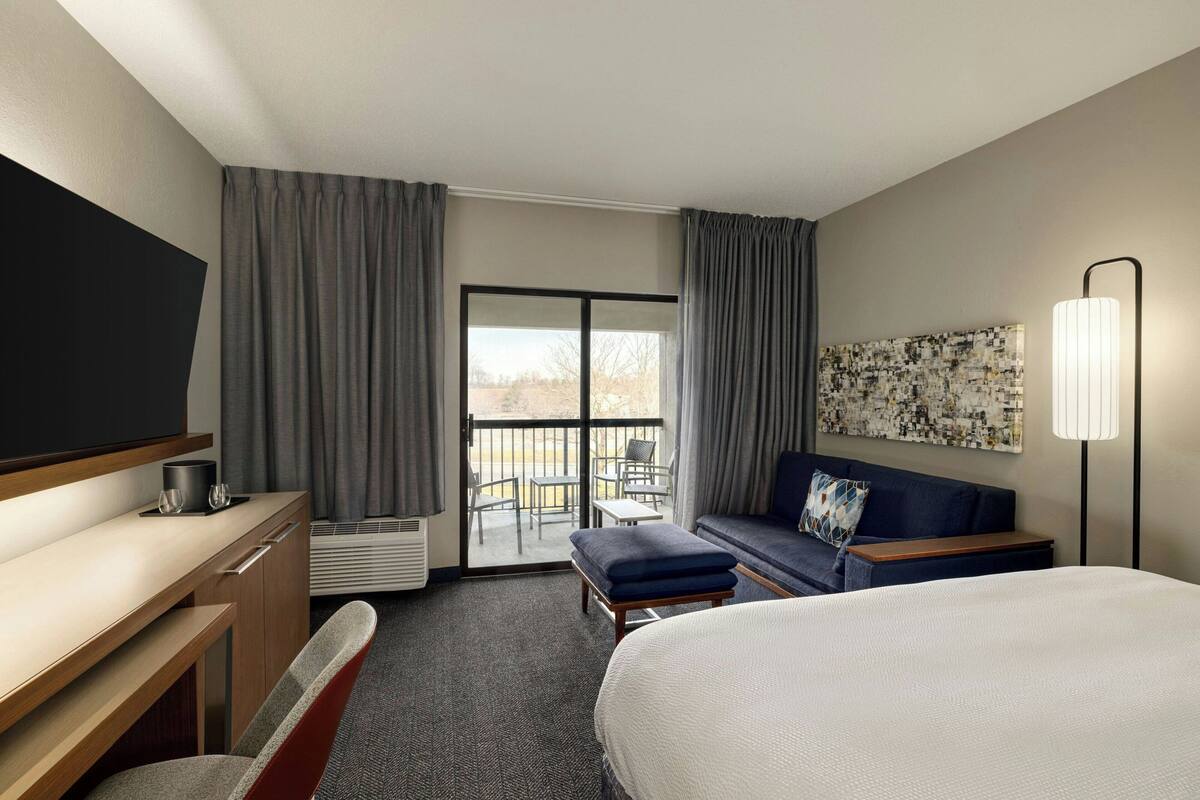 Ein Hotelzimmer mit Bett, Schreibtisch, Stuhl, Sofa, Fernseher, Lampe und einem Balkon mit Sitzgelegenheiten.