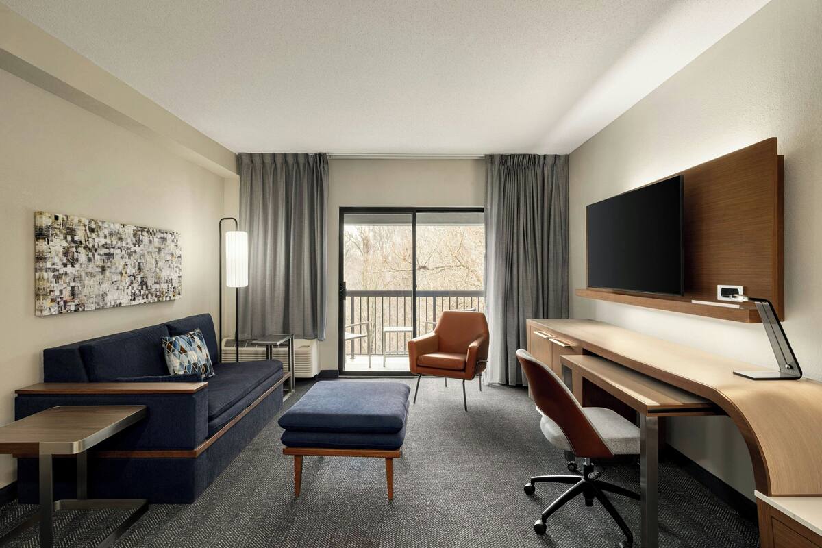 Ein Hotelzimmer mit Sofa, Hocker, Schreibtisch, Stuhl, Fernseher und einem Balkon mit Vorhängen.