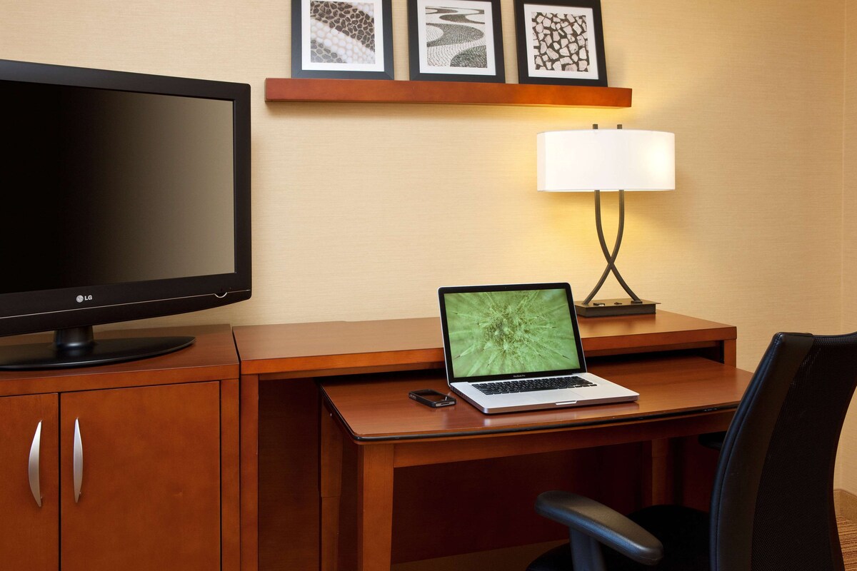 A hotel room with a wooden desk, a laptop, a flat-screen TV, a lamp, and a chair.