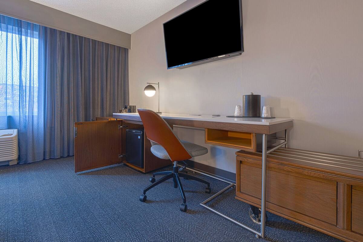 A hotel room with a desk, chair, television, lamp, and a view of the outdoors.