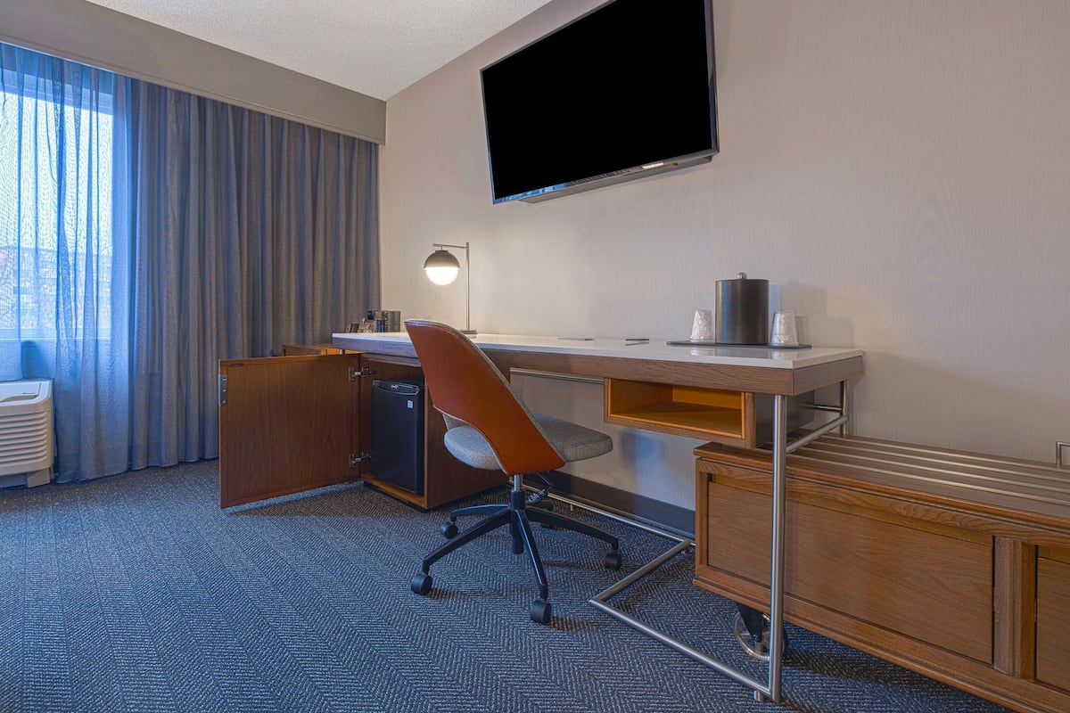 Habitación de hotel con escritorio, silla, televisión, lámpara y vistas al exterior.