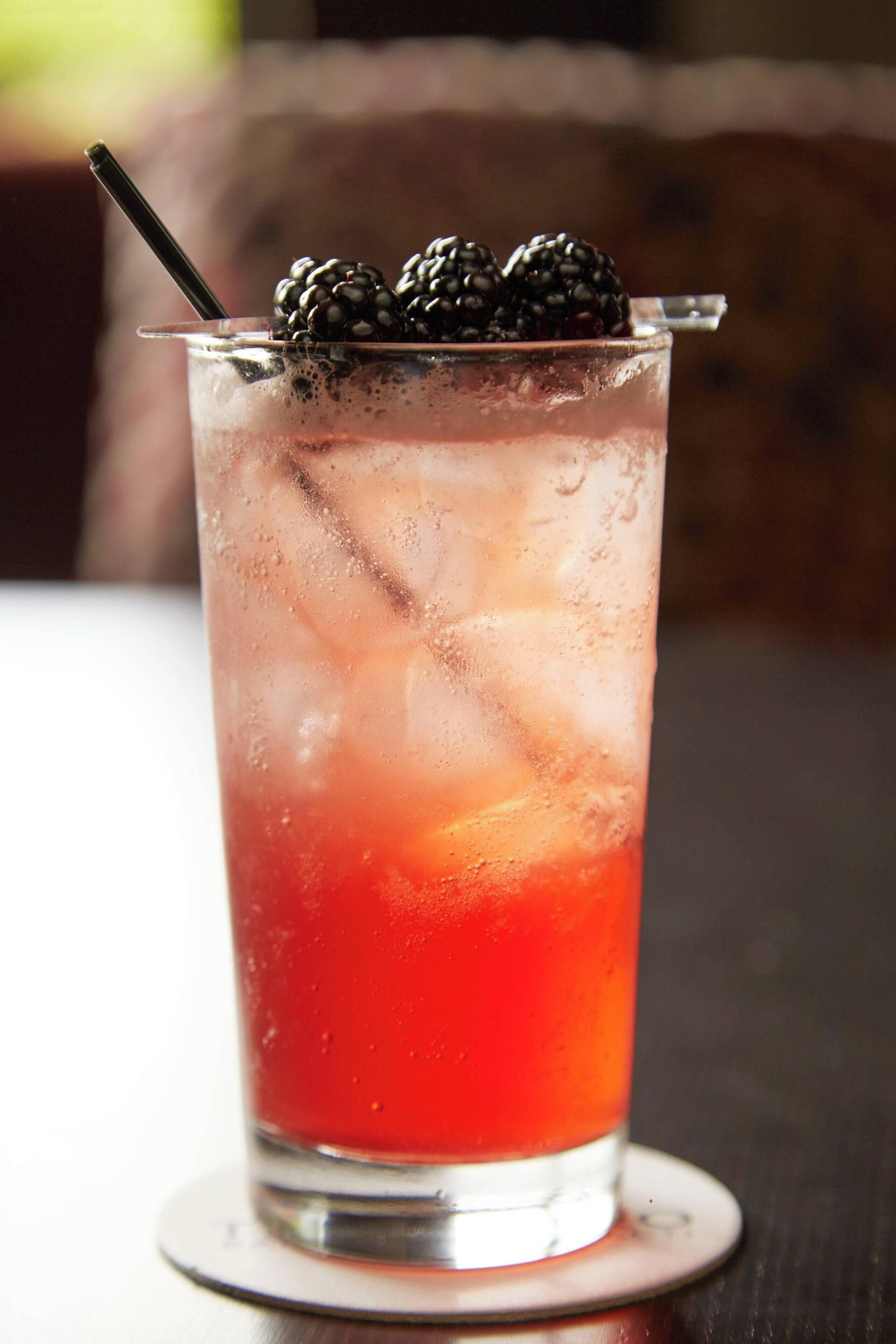 A tall glass with a red beverage, ice cubes, and blackberries on a coaster.