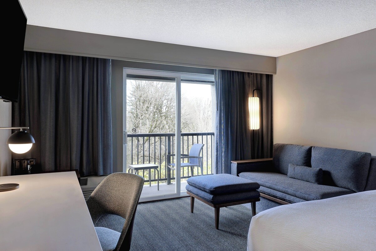A hotel room with a sofa, a chair, a small table, and a desk. There is a balcony with a chair and a table outside.