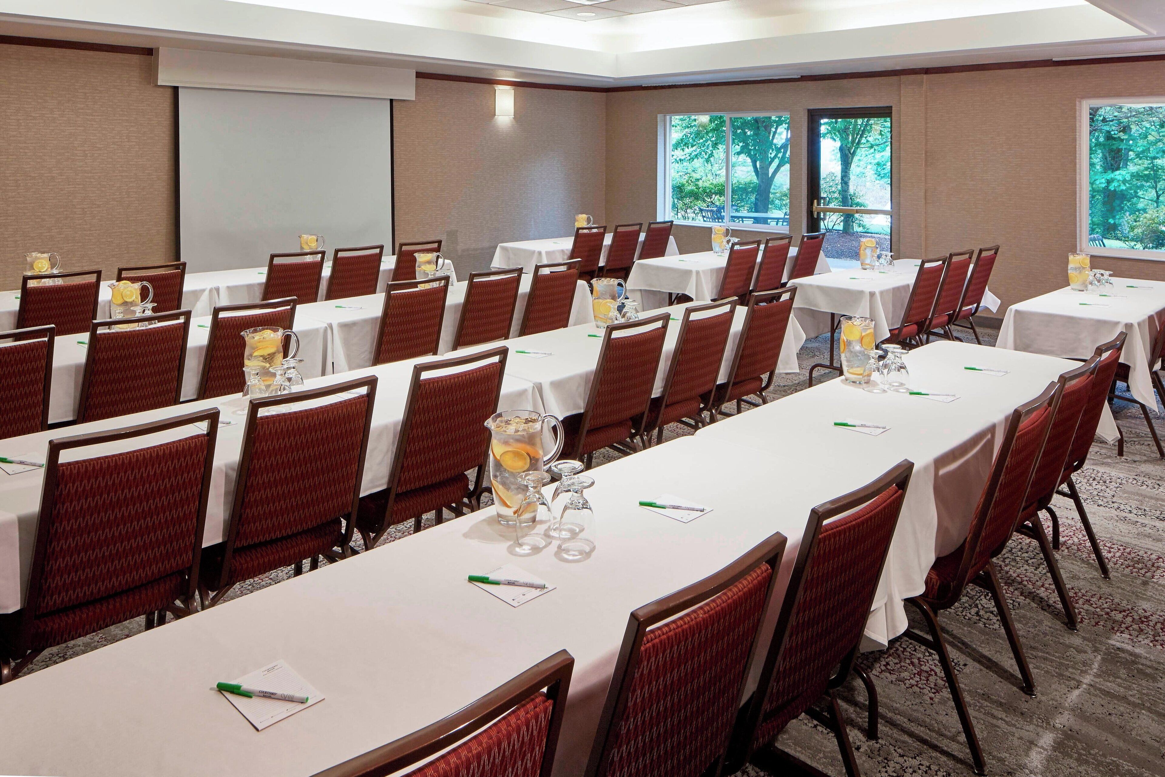 A conference room with multiple tables and chairs, equipped with water pitchers and glasses, and a projector screen on the wall.