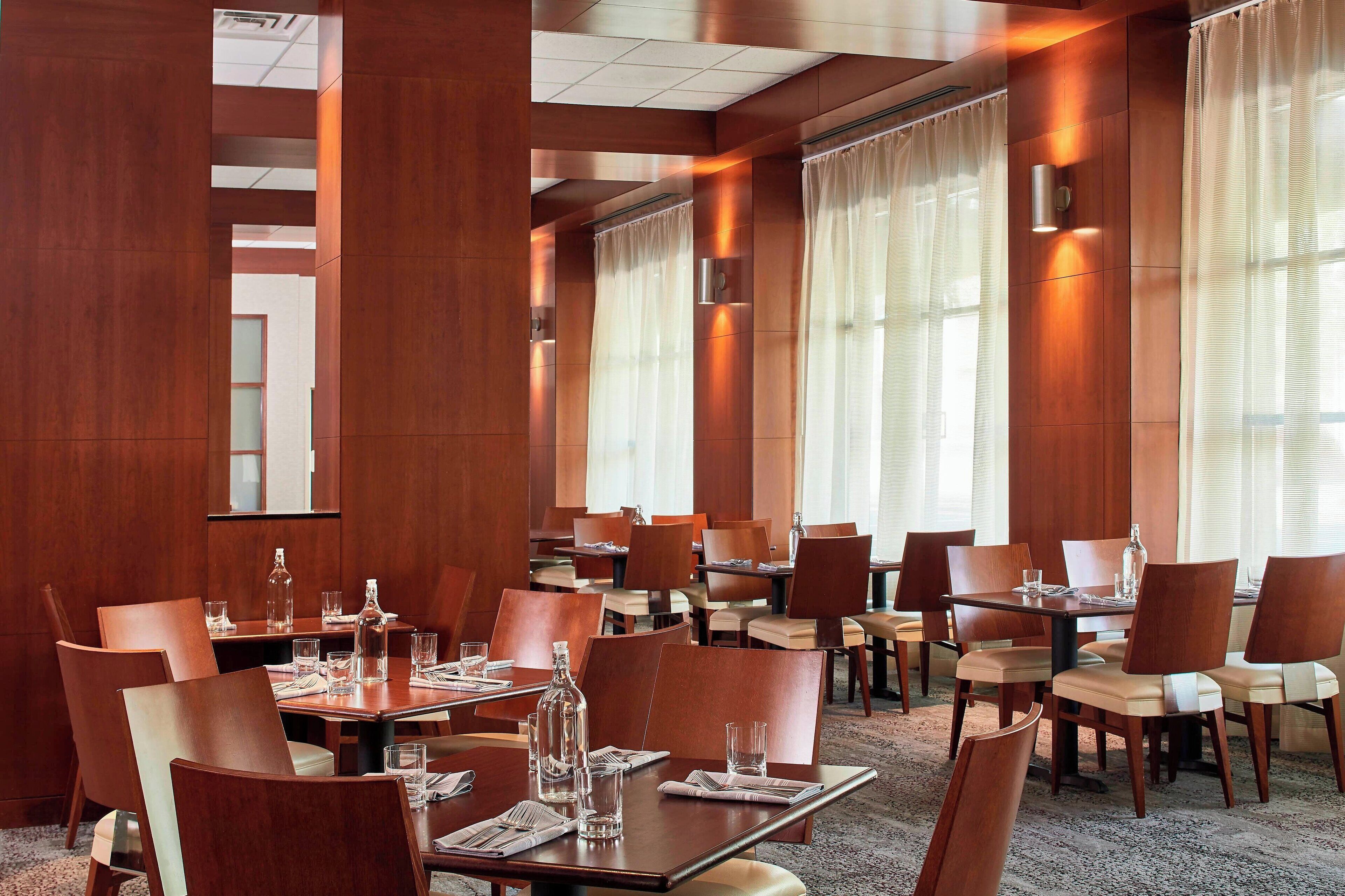 Un espace de restauration d’hôtel comprenant plusieurs tables et chaises, de grandes fenêtres avec des rideaux et un aménagement intérieur moderne.