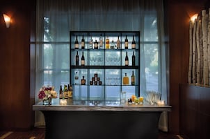 Zona de bar con estantes llenos de botellas y copas, barra y ventana con cortinas.