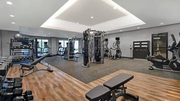 A modern gym with various exercise equipment, a wooden floor, and a large window providing natural light.