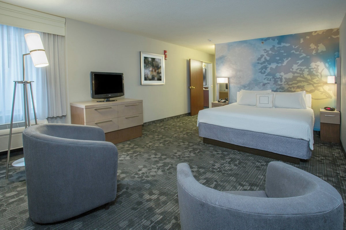 A hotel room with a bed, two armchairs, a desk with a television, a lamp, and a painting on the wall.