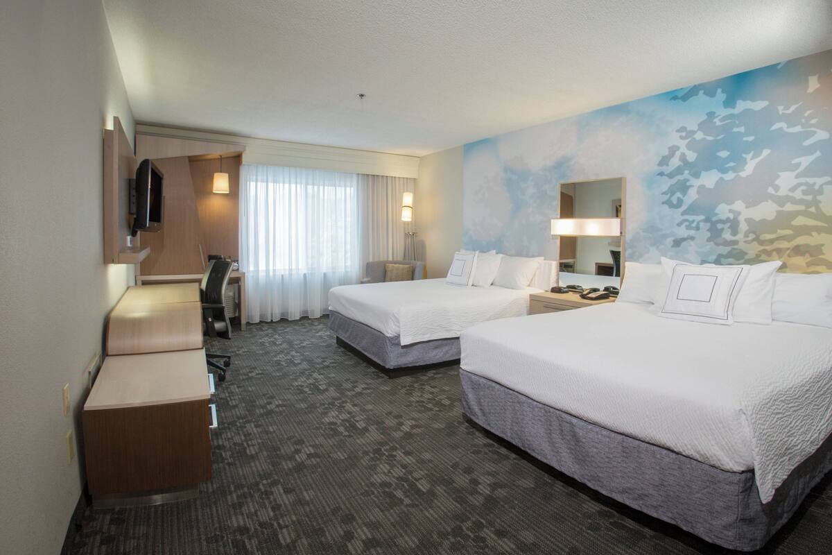 A hotel room with two beds, a desk, a chair, a TV, and a large window with curtains.