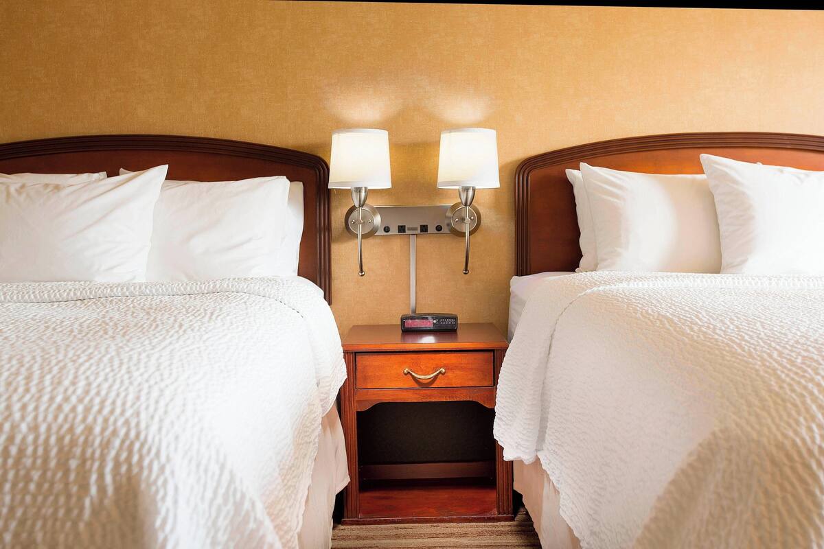 A hotel room with two beds, a nightstand with a clock, and a wall-mounted lamp.
