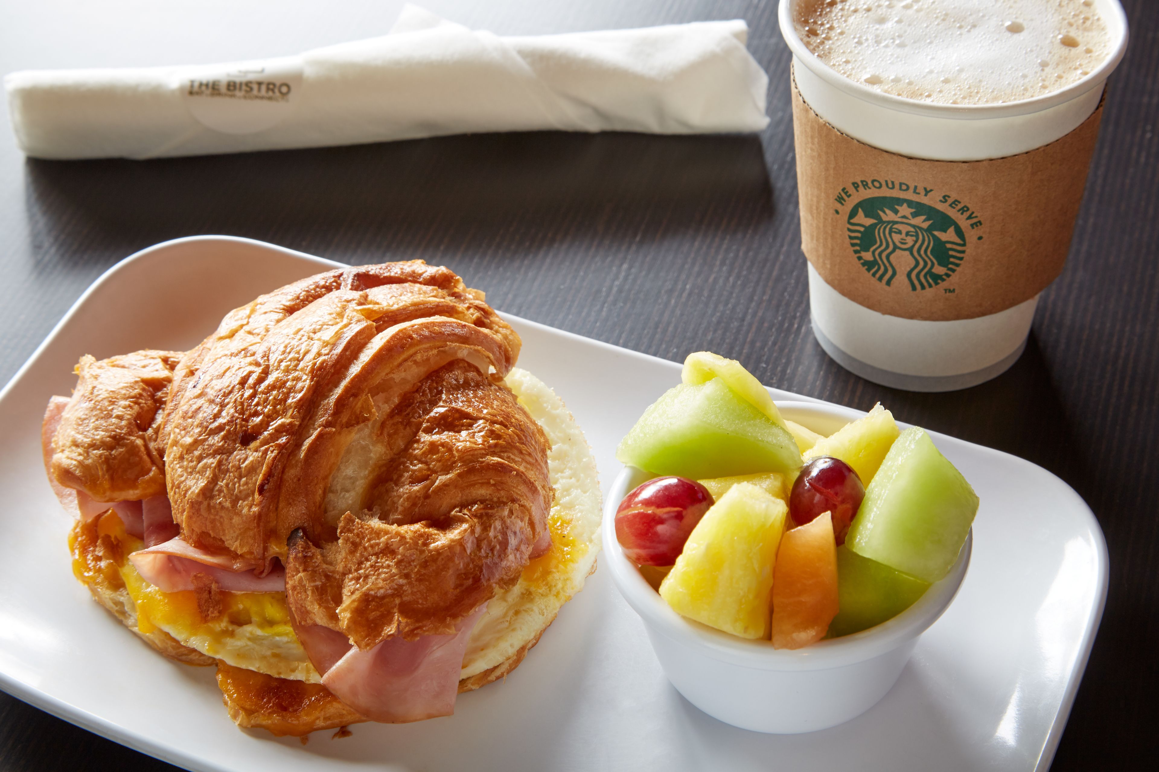 Un sándwich de croissant con jamón y queso, una taza de café y un bol de fruta sobre una bandeja.