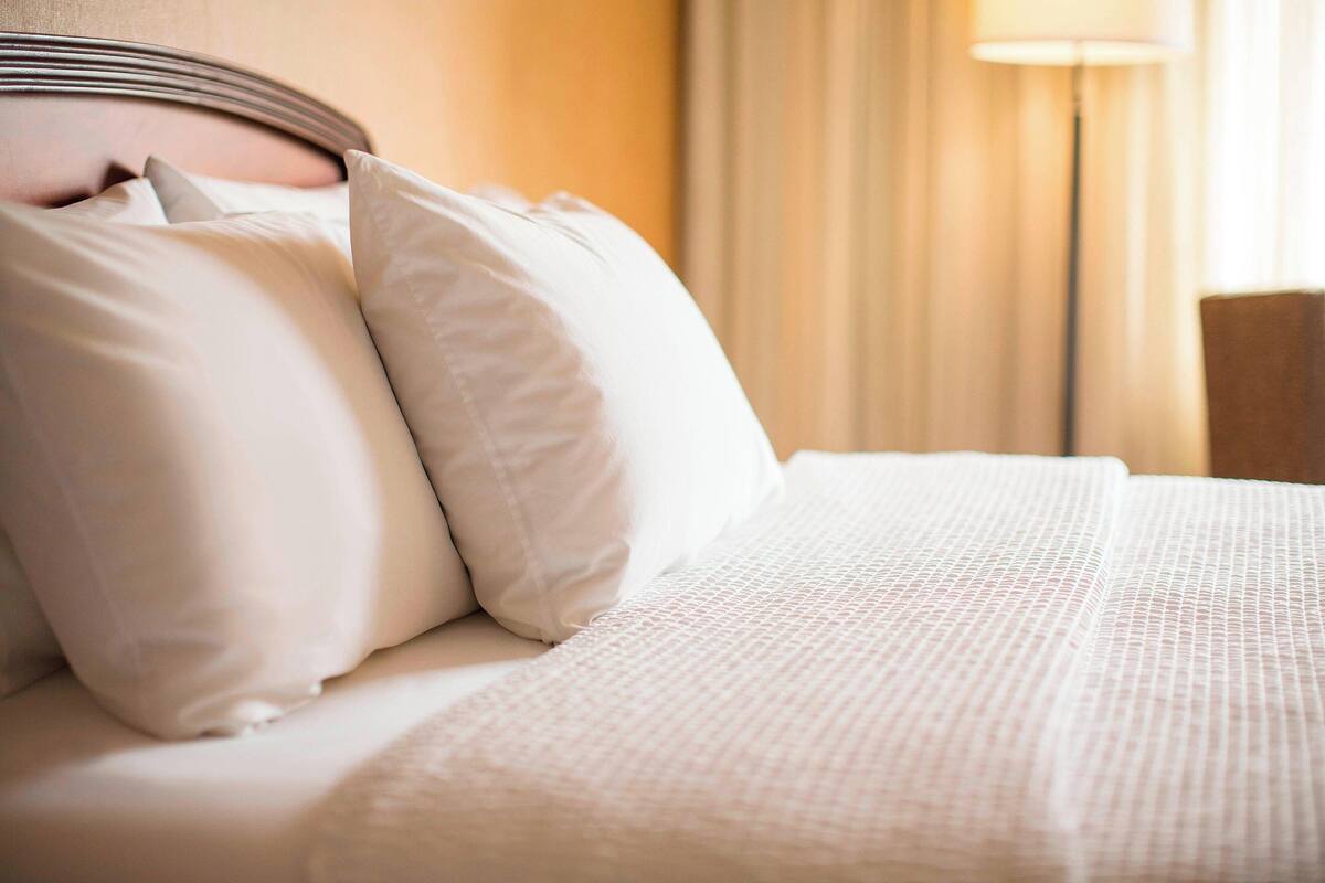 A neatly made bed with white linens and pillows, a headboard, and a lamp in the background.