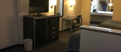 A hotel room with a bed, a desk, a chair, a television, and a small trash bin.