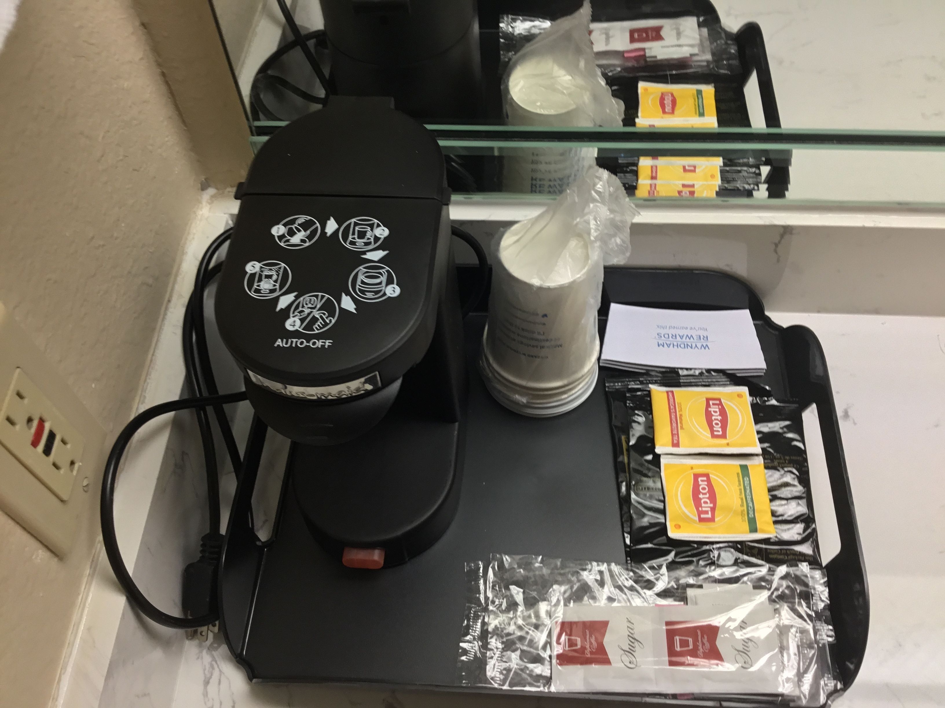 Máquina de café com função de desligamento automático, jarra de café e chá Lipton embalado, juntamente com açúcar, sobre um balcão.