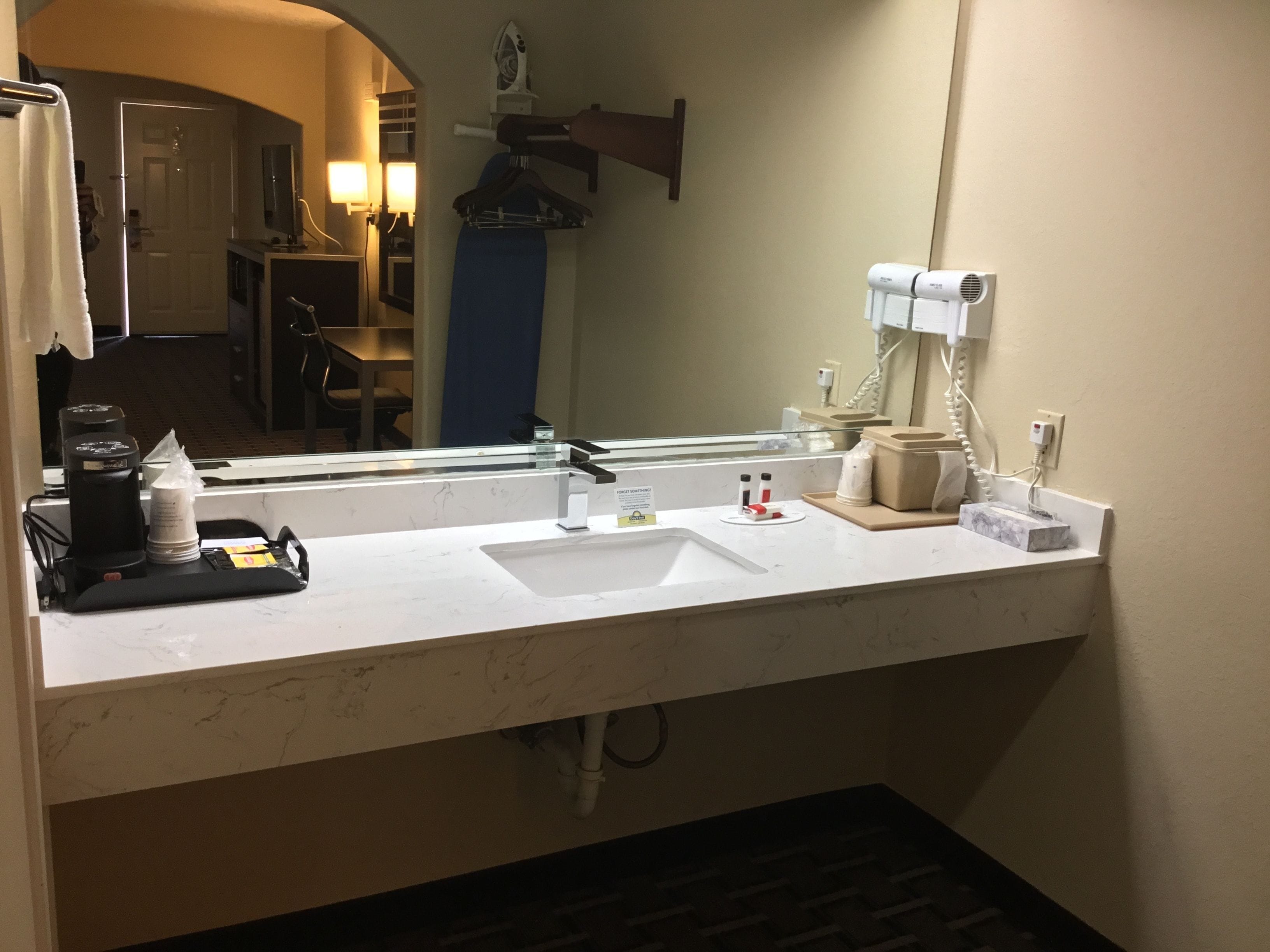 Une salle de bain d’hôtel avec un lavabo blanc, un miroir, un sèche-cheveux et une machine à café.