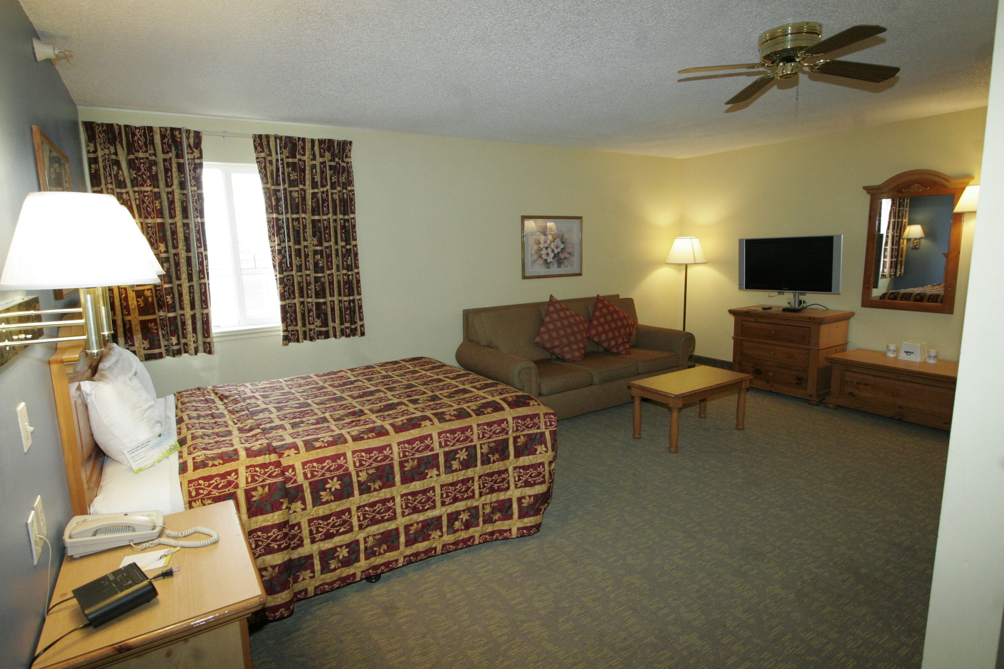 A hotel room with a bed, a sofa, a desk with a lamp, a television, a dresser, and a ceiling fan.