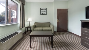 A hotel room with a window, a sofa, a table, a lamp, a door, and a TV. - Days Inn & Suites by Wyndham Dallas (Dallas)