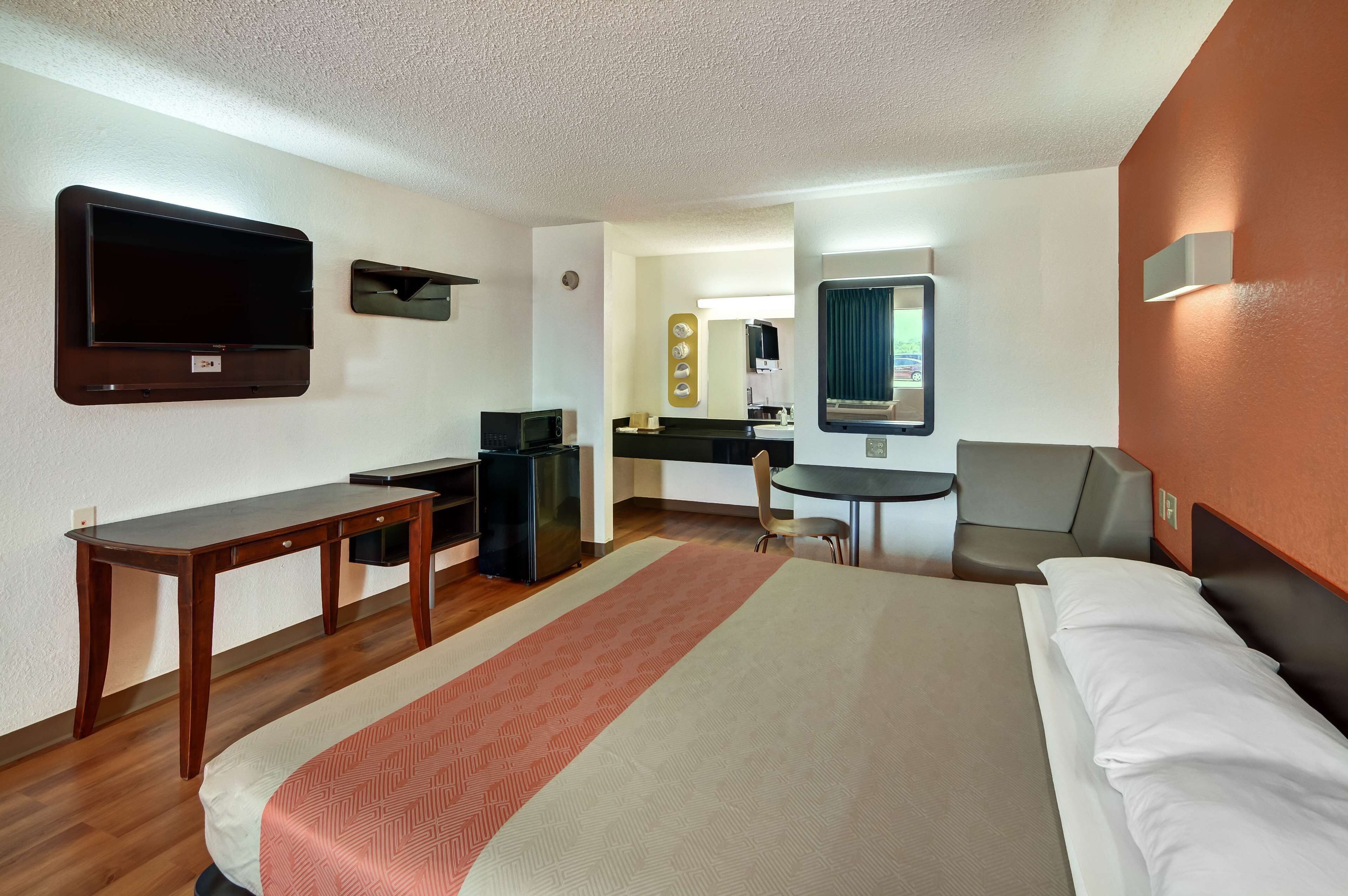 Quarto de hotel com cama, escrivaninha, cadeira, televisão, pequena cozinha e espelho de banheiro.